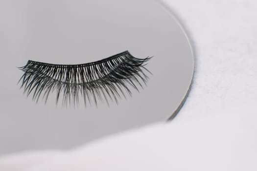 Black and long artificial eyelashes using for lash extension procedure lying on round mirror on white table. Isolated fake cilia reflecting in looking-glass, making image of fluffiness and volume.