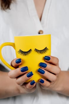Yellow cup of beverage with black fake eyelashes sticked on outside in hands of unrecognizable lady with blue manicure. Lashmaker holding mug with material for lash extension procedure. Vertical.