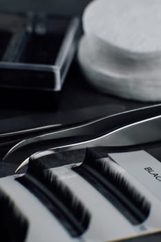 Close up of black artificial eyelashes in palette lying close to isolated tweezers on table prepared for lash extension procedure. Different tools and consumable for creating flawless look. Vertical.
