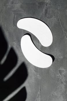 Pair of white patches using as insulation of eyelashes and applying on lower eyelids, lying on gray table with cast shadow of leaf near. Isolated consumable for lash extension procedure. Vertical.