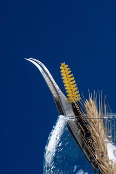 Close up of tweezers and yellow brush using for eyelash extension procedure left in wineglass standing against dark blue background. Different tools in wet goblet with branch of wheat. Vertical.