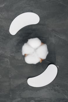 View from above of pair of white patches, applying on lower eyelids and isolating lashes, using for eyelash extension procedure lying out on gray table around cotton pads in form of flower. Vertical.