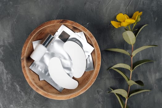 View from above of disposable patches for eyelash extension procedure lying in wooden bowl full of materials on gray table near yellow flower. White consumable for isolating lashes in round plate.