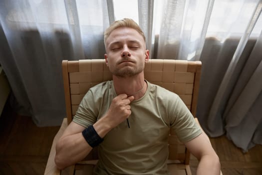 Depressed lonely man soldier with emotional problem and war syndrome feeling nervous, suffering from PTSD touching his badge tag sitting on chair alone at home