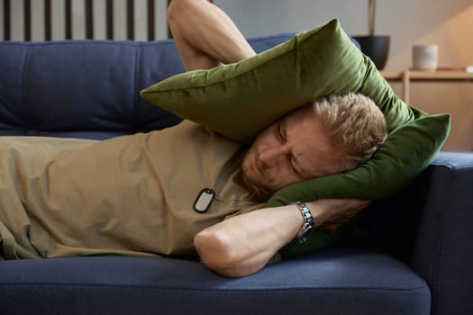 Stressed soldier man covering ears with pillow trying drown out voices, sounds of explosions and gunshots in his head. PTSD symptom