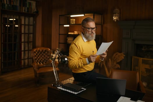 Busy focused mature man reading paper report while working at home office in evening. Senior male doing paperwork checking bills accounting financial income sitting on desk with pc laptop computer