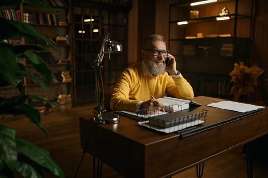 Happy elderly man laughing and talking mobile phone while working at home office in evening
