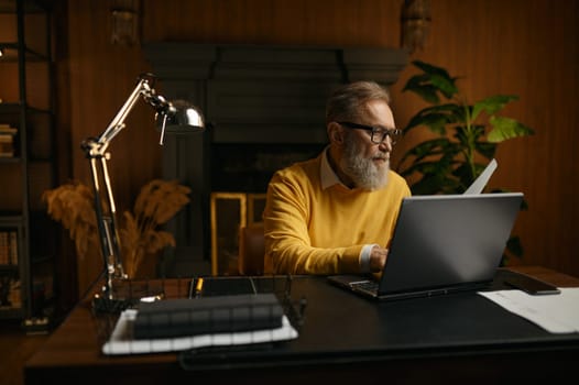 Senior man typing on laptop keyboard while sitting at table and working at home office in evening