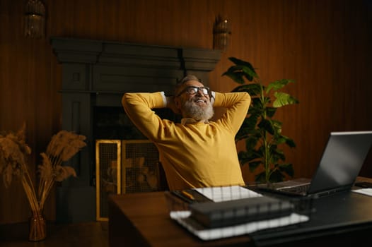 Excited mature man feeling satisfaction relaxed sitting at table with laptop in home office. Happy dreamy senior male holding hands behind head looking up with smile on face think of future success