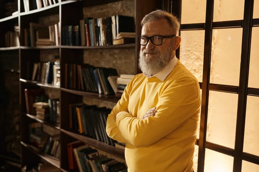 Pensive gray-haired senior man standing with arms crossed on chest at home library aristocratic interior. Mature older man leaned at bookshelves