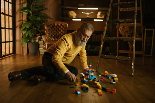 Senior old man playing toy building blocks while sitting on home floor during cozy evening. Handsome adult businessman on retirement