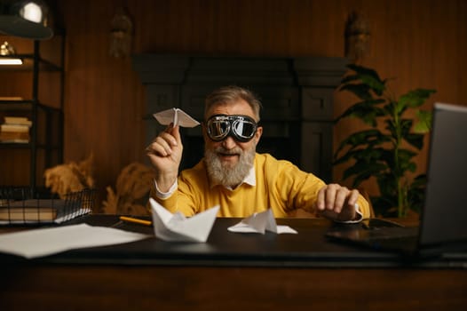 Funny overjoyed mature man wearing pilot goggles playing with origami plane while sitting at workplace in home office in evening