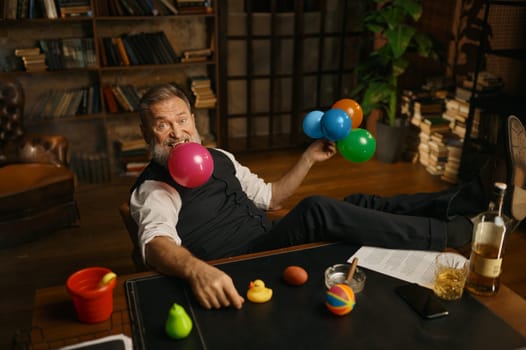 Portrait of senior business man blowing up balloon for success celebration. Grey-haired fashioned mature male boss sitting at work table in home office