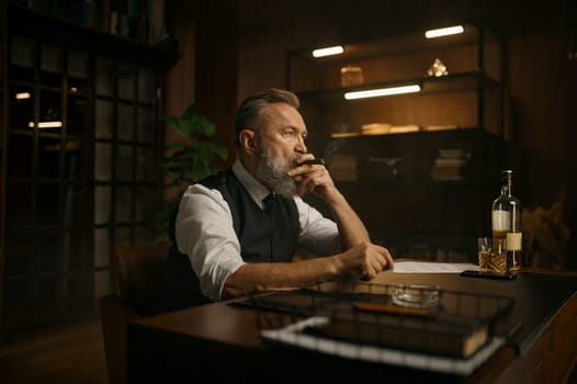 Pensive overwhelmed senior business man smoking cigar while sitting at work table with opened drink of alcohol in home office. Bad thoughts, professional burnout concept