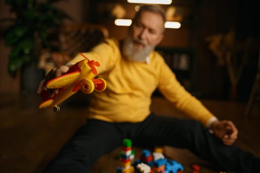 Selective focus of elderly man in trendy fashion outfit playing toy airplane and building blocks while sitting on home floor. Work break, mental health, dreaming about future travel concept