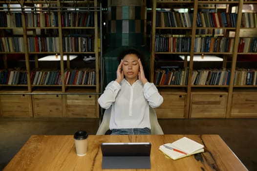 Tired overworked female office worker touching head massaging temples suffering from headache or tension while sitting at desk table front of computer