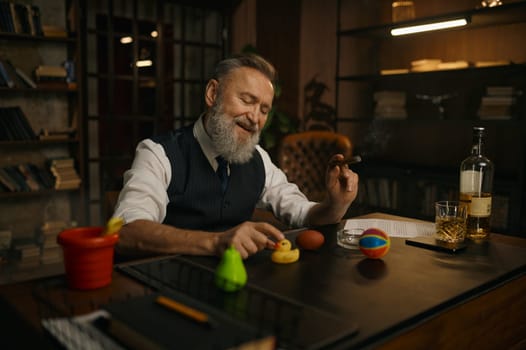 Funny elegant older businessman smoking cigar and playing toys at work table in home office