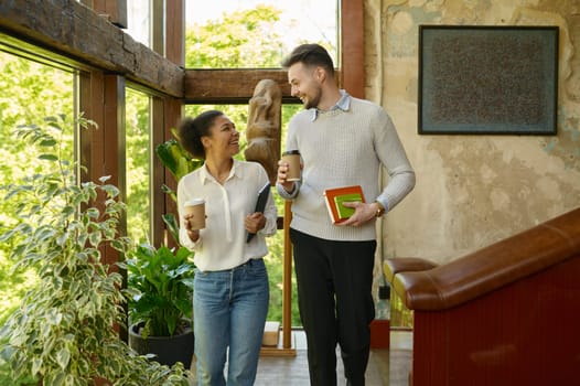 Diverse multiethnic man and woman colleagues taking coffee break talking and walking among hallway in eco-friendly office
