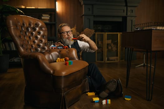 Funny senior businessman playing toys enjoying fun time after hard working day in home office. Elderly male sitting on floor nearby armchair