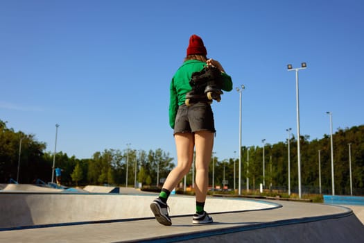 Casual girl carrying rollerblades in hands walking to training class. Outdoor rollerdrome for practicing freestyle speed riding