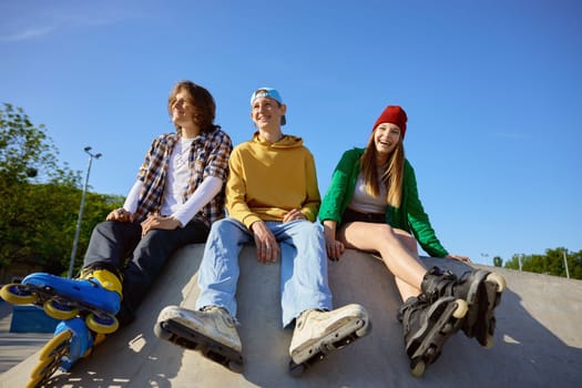 Joyful rollerskaters team of teenage people sitting on ramp in skate park. Extreme sports training and processional equipment for teens