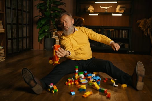 Selective focus of elderly man in trendy fashion outfit playing toy airplane and building blocks while sitting on home floor. Work break, mental health, dreaming about future travel concept