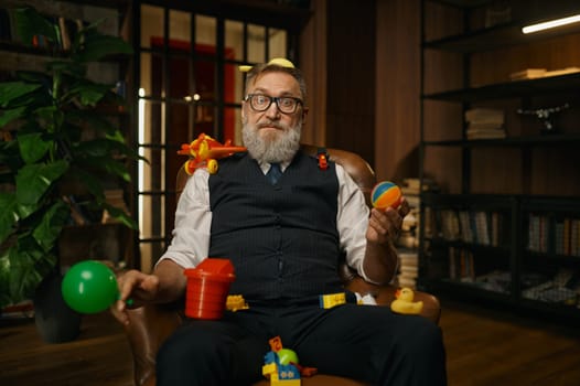 Portrait of rich senior businessman sitting in leather armchair with toys looking at camera over cozy home office interior