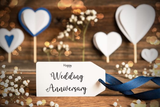 Label With English Text Happy Wedding Anniversary. White Festive And Atmospheric Decoration Like Hearts, Flowers And A Blue Bow. Vintage, Wooden Background With Sparkling Light.