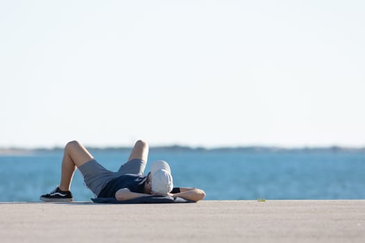 A man skater in sportswear rests at the waterfront lying on his back. Mid shot