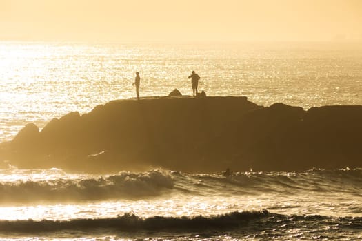 Silhouettes of people standing on a cliff in the middle of the sea and fishing at sunset. Mid shot