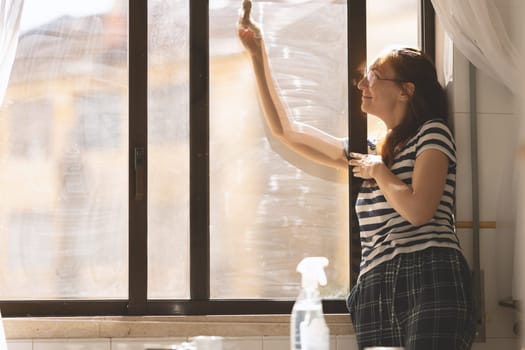Smiling adult woman housewife washing the outside of a window in the kitchen. Mid shot