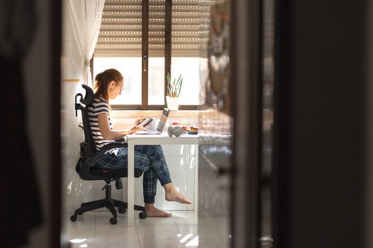 An adult woman freelancer working at her laptop at the kitchen table holding a phone. Mid shot