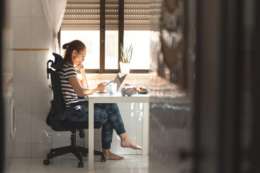 Thoughtful woman freelancer working at her laptop at the kitchen table holding a phone. Mid shot