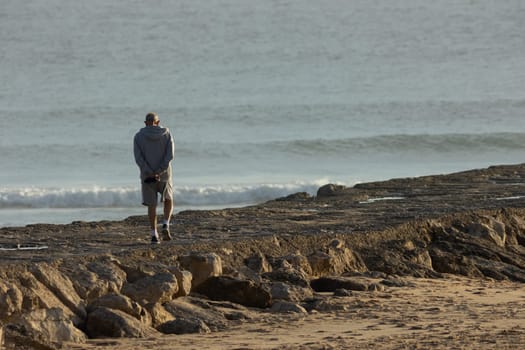 A lonely elderly man walks on a rocky ledge by the sea. Mid shot