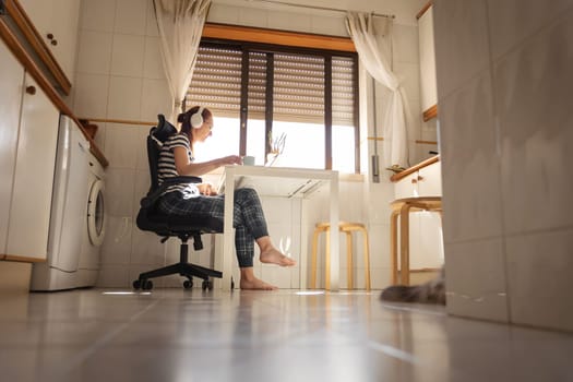 An adult woman freelancer in headphones working at the kitchen. Mid shot
