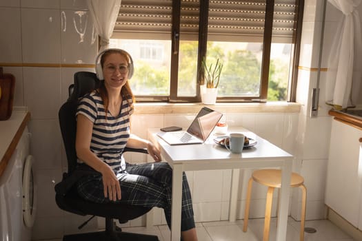Smiling adult woman freelancer sitting by the kitchen table. Mid shot