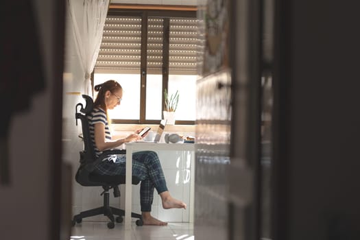 An adult woman working at her laptop at the kitchen table holding a phone. Mid shot