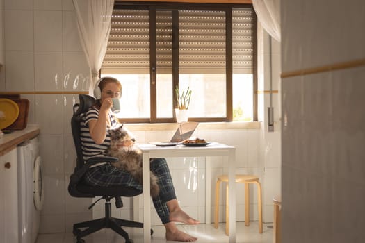 A woman drinking coffee sitting at the kitchen table with her cat on her laps. Mid shot