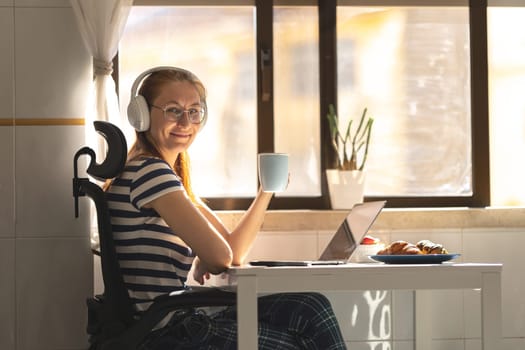 Smiling adult woman freelancer in headphones sitting by the kitchen table with a cup of coffee. Mid shot