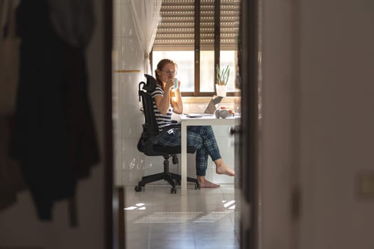 Adult woman freelancer working at the kitchen table holding a cup of coffee - looking in the camera. Mid shot