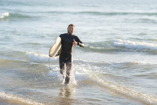 Smiling man surfer running out of the sea holding a surfboard and showing shaka. Mid shot