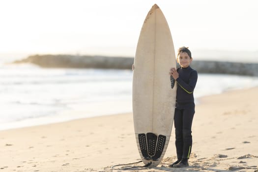 A little boy in a wetsuit holding a big white surfboard standing on the seashore. Mid shot