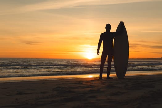 A fit man standing on the seashore leaning on a surfboard at sunset. Mid shot
