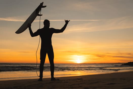 Silhouette of an athletic man standing on the seashore holding a surfboard and showing shaka at sunset. Mid shot