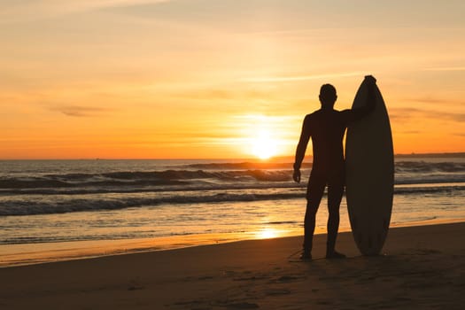 A fit man stands on the seashore leaning on a surfboard at sunset. Mid shot