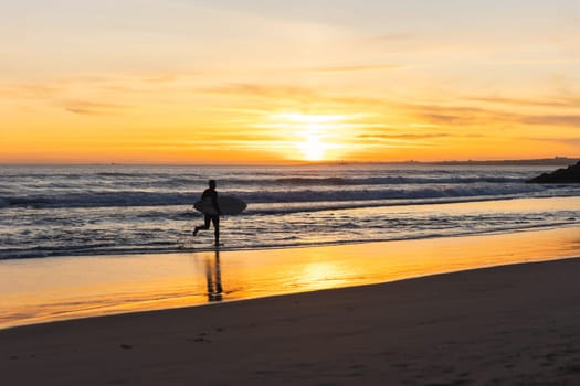 A man surfer running on the seashore at sunset. Mid shot
