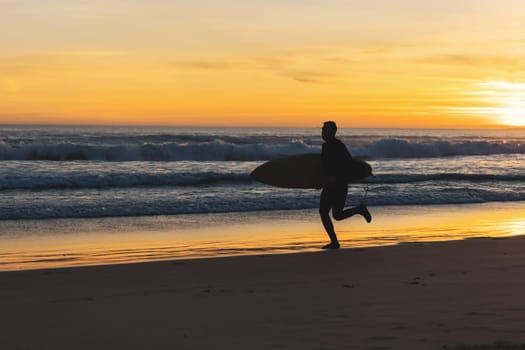 A man surfer running on the seashore holding a surfboard at sunset. Mid shot