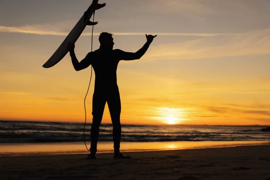 Silhouette of man standing on the seashore holding a surfboard and showing shaka at sunset. Mid shot
