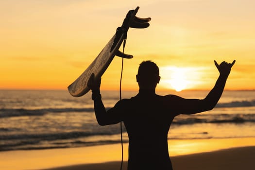 Silhouette of an athletic man holding a surfboard showing shaka at sunset. Mid shot