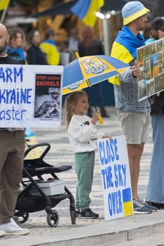 10-29-2022 LISBON, PORTUGAL: Ukrainian protest in Lisbon - little girl under a blue-yellow umbrella holds a poster CLOSE THE SKY OVER UKRAINE. Mid shot